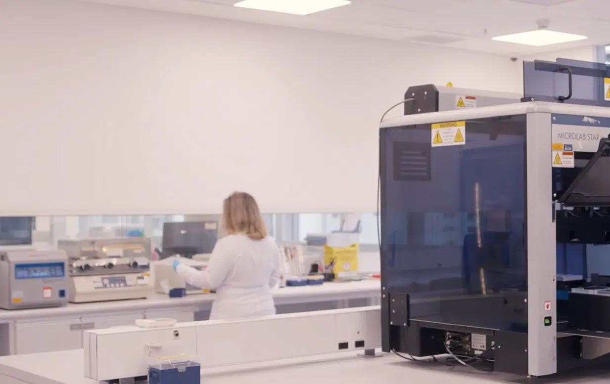 Ambiente laboratorial moderno com pesquisadora ao fundo manuseando equipamentos de análise genética