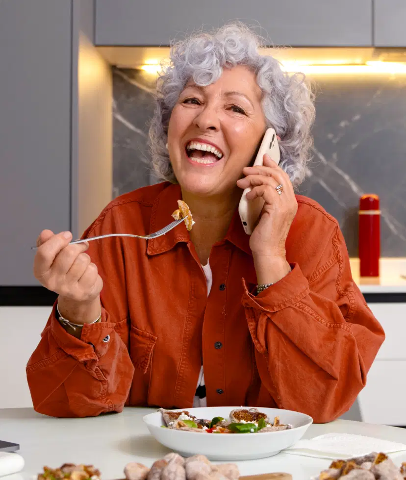 Mulher de meia idade sorridente, comendo uma salada enquanto fala ao celular em uma cozinha moderna